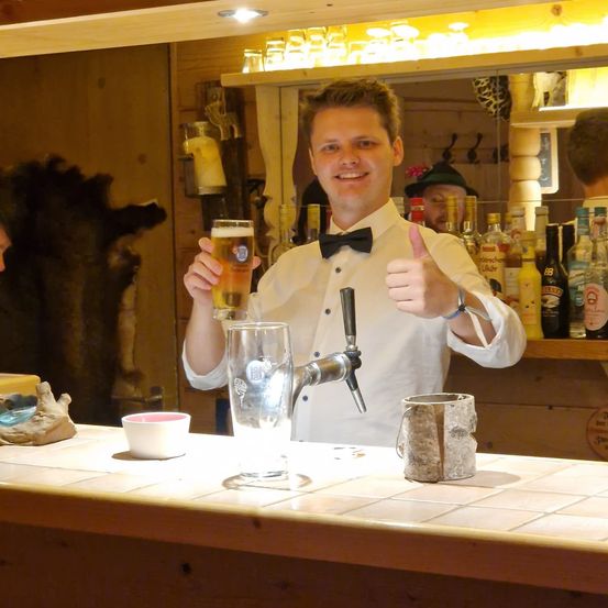 Ein Mann mit Fliege lächelt in die Kamera, während er ein Bierglas in einer rustikalen Bar hält.