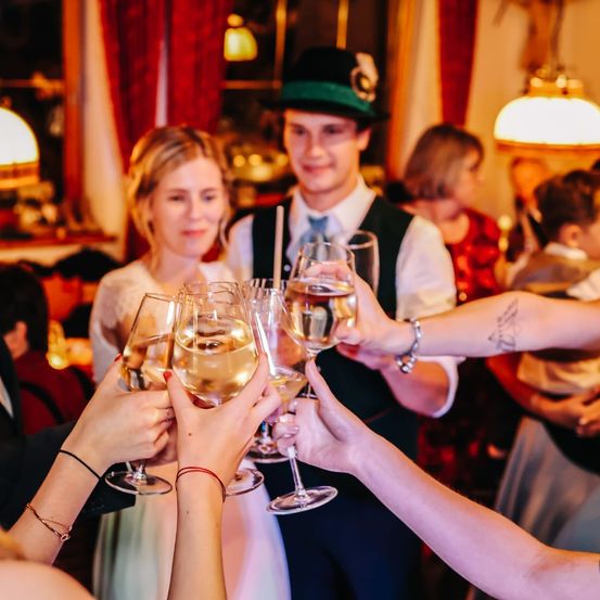 Mehrere Personen halten Weinglas in einem Raum. Eine Frau in einem weißen Kleid lächelt. Ein Mann trägt einen Hut und eine Krawatte. Eine Lampe ist an der Wand. Ein Junge steht in der Nähe einer Frau in einem roten Kleid.