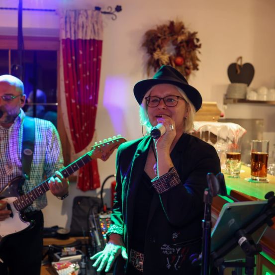 Eine Frau singt mit einer grünen Lampe in ein Mikrofon, während ein Mann Gitarre spielt. Die Szene spielt in einem Raum mit einer dekorierten Wand und verschiedenen Gegenständen wie einem Kranz, Herz und Gläsern.