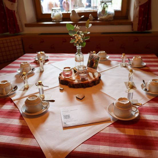 Ein Esstisch für vier Personen mit Tassen, Untertassen und kleinen Flaschen. Ein holzner Zentrumsstück hält eine Vase mit Blumen, ein Foto und kleinen Gläsern. Ein weißer Stoff bedeckt den Tisch.