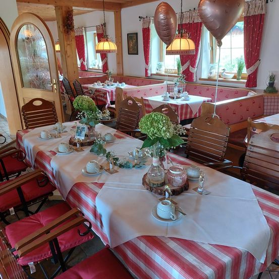 Ein Esszimmer mit mehreren Tischen und Stühlen, jeder mit Tassen, Untertassen und Teekannen besetzt. Die Tische sind mit weiß-rot gestreiften Tischdecken bedeckt. Der Raum hat eine rustikale Holzdekoration, mit Luftballons und einem gerahmten Bild auf den Tischen.