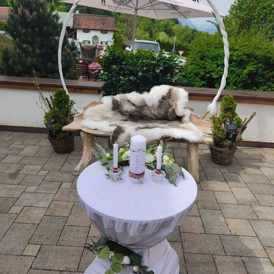 Ein Hochzeitsaltar auf einer Terrasse mit einem Tisch, drei Kerzen, Blumen und einem Fell auf einer Bank unter einem Schirm. Die Umgebung besteht aus Pflanzen, einem Holzzaun und einem Haus im Hintergrund.