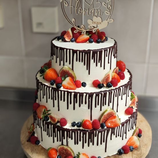 Dreischichtiger Kuchen mit weißer Glasur, Schokoladen-Drip und frischen Früchten. Die oberste Schicht hat ein herzförmiges Schild mit Florian 12.10.2024. Auf einem Holzuntergrund platziert.