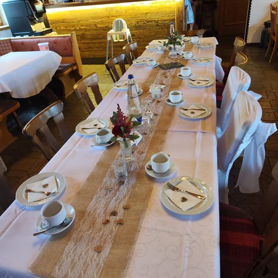 Ein langer Tisch, der für eine formelle Veranstaltung gedeckt ist, mit weißer Tischdecke, Tassen, Untertassen und Besteck. Ein dekorativer Tischläufer und Blumen sind ebenfalls vorhanden. Die Stühle sind mit weißem Stoff bezogen.