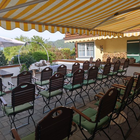 Reihen von Stühlen unter einem gestreiften Vordach, wahrscheinlich für eine Veranstaltung im Freien, mit einer Kulisse aus Grün und Bergen.