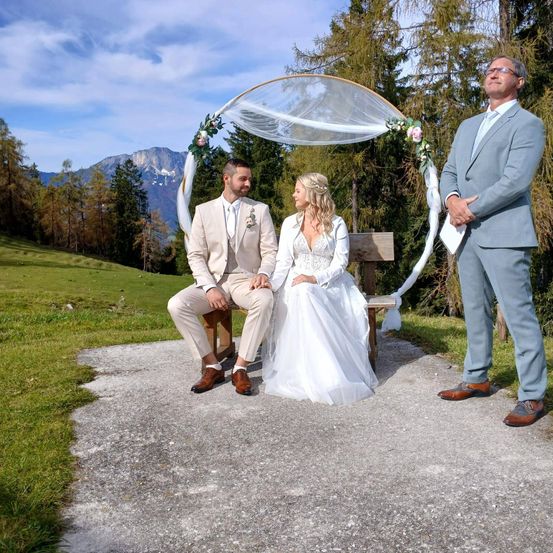 Eine Braut und ein Bräutigam sitzen auf einer Bank unter einem Blumenbogen, mit einem Mann in Anzug hinter ihnen. Sie sind von einem grünen Feld und Bergen umgeben.