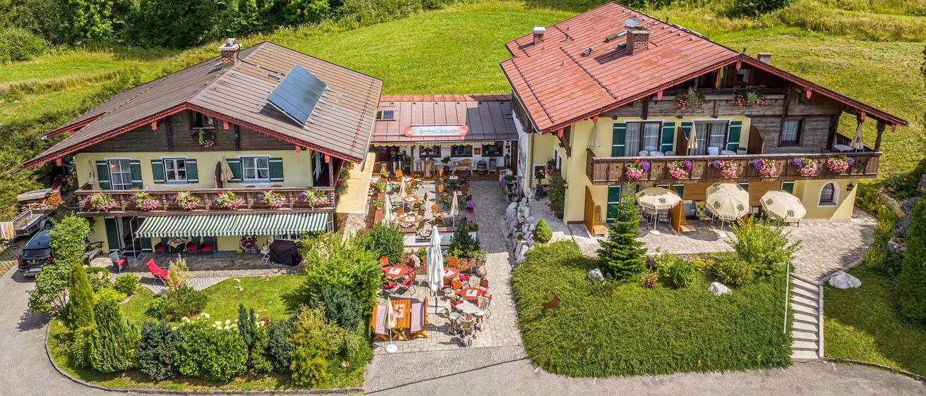 Ein Luftbild eines ländlichen Resorts mit zwei Häusern, einem Restaurant und Sitzgelegenheiten im Freien. Die Häuser haben rote Dächer und Solarpanels. Bäume und Berge sind im Hintergrund.