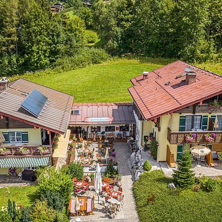 Luftaufnahme eines rustikalen gelben Hauses mit rotem Dach und Solarpanels, umgeben von üppiger Grünfläche, mit einem Essbereich im Freien davor.