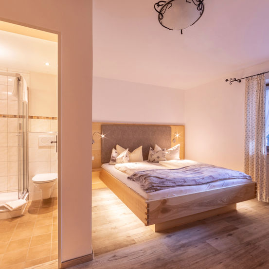 Ein Schlafzimmer mit Bett, Kissen und einem Fenster mit Vorhängen. Das Badezimmer hat eine Toilette, Dusche und ein Handtuch an der Wand.