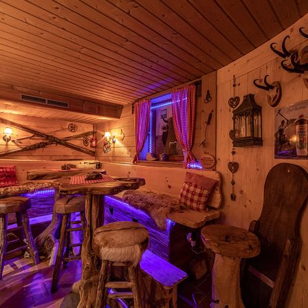 Ein rustikaler Loungebereich mit Holzmöbeln und lila Beleuchtung. Es gibt zwei Tische mit Hockern, eine Bank mit Kissen und an der Wand befestigte Geweihe. Der Raum hat auch ein Fenster mit einem roten Vorhang und einer Lampe.