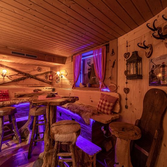 Ein rustikaler Loungebereich mit Holzmöbeln und lila Beleuchtung. Es gibt zwei Tische mit Hockern, eine Bank mit Kissen und an der Wand befestigte Geweihe. Der Raum hat auch ein Fenster mit einem roten Vorhang und einer Lampe.