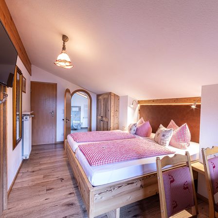 Ein gemütliches Schlafzimmer mit Holzboden und -wänden. Es verfügt über ein Doppelbett mit rot-weiß kariertem Bettzeug, zwei Holzstühlen und einem montierten Fernseher. Das Zimmer hat auch einen Holzschrank und einen Spiegel.