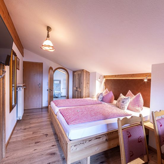 Ein gemütliches Schlafzimmer mit Holzboden und -wänden. Es verfügt über ein Doppelbett mit rot-weiß kariertem Bettzeug, zwei Holzstühlen und einem montierten Fernseher. Das Zimmer hat auch einen Holzschrank und einen Spiegel.