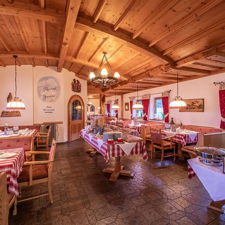 Ein Restaurant mit Holzdecke und Wänden. Es hat Tische mit rot-weiß karierten Tischdecken. Es gibt Buffettische mit Essen. Die Stühle sind aus Holz. Es hängen Lampen an der Decke. An der Wand befindet sich ein Schild.