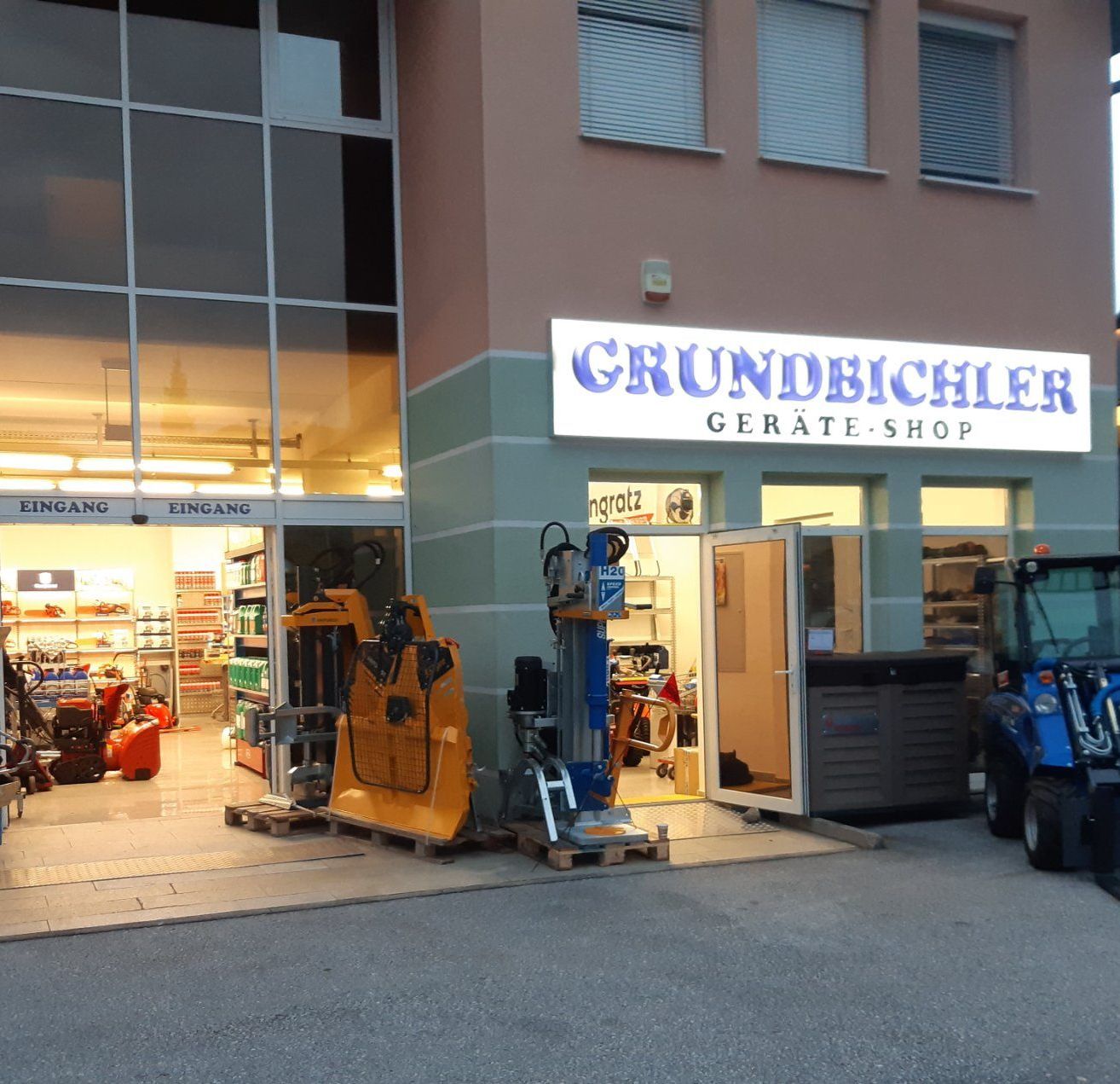 Ein Geschäft mit dem Schild 'Grundbiehler Gerate-Shop'. Das Geschäft hat eine Glaswand und Eingang, mit schweren Maschinen draußen ausgestellt. Es steht ein Gabelstapler auf der rechten Seite.