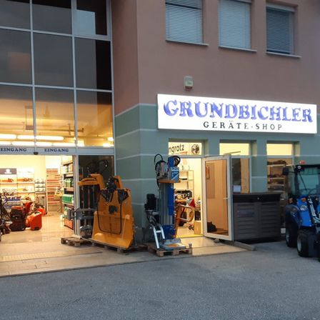 Ein Geschäft mit dem Schild 'Grundbiehler Gerate-Shop'. Das Geschäft hat eine Glaswand und Eingang, mit schweren Maschinen draußen ausgestellt. Es steht ein Gabelstapler auf der rechten Seite.