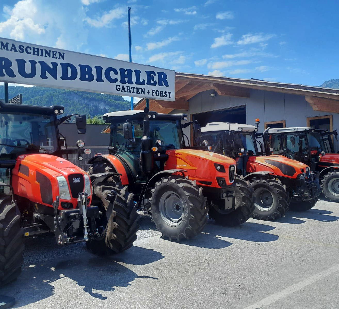 Mehrere orange Traktoren sind vor einem Gebäude mit dem Schild Maschinen Grundbichler Garten + Forst geparkt.