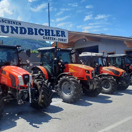Mehrere orange Traktoren sind vor einem Gebäude mit dem Schild Maschinen Grundbichler Garten + Forst geparkt.