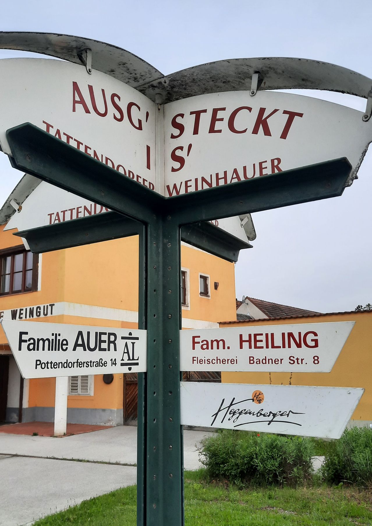Ein Wegweiser mit Wegbeschreibungen zu verschiedenen lokalen Unternehmen. Oben links steht 'AUSG', oben rechts 'STECKT S', unten links 'Familie AUER AL' und unten rechts 'Fam. HEILING'.