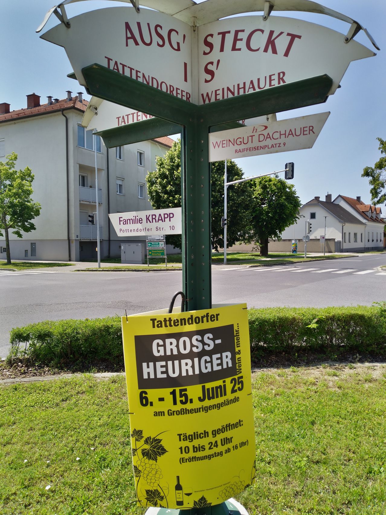 Ein gelbes Schild mit 'Gross-Heuriger 6.-15. Juni 25' steht neben einer Straße mit Gebäuden und Bäumen. Straßenschilder zeigen nach Tattendorfer und Weingut Dachauer.