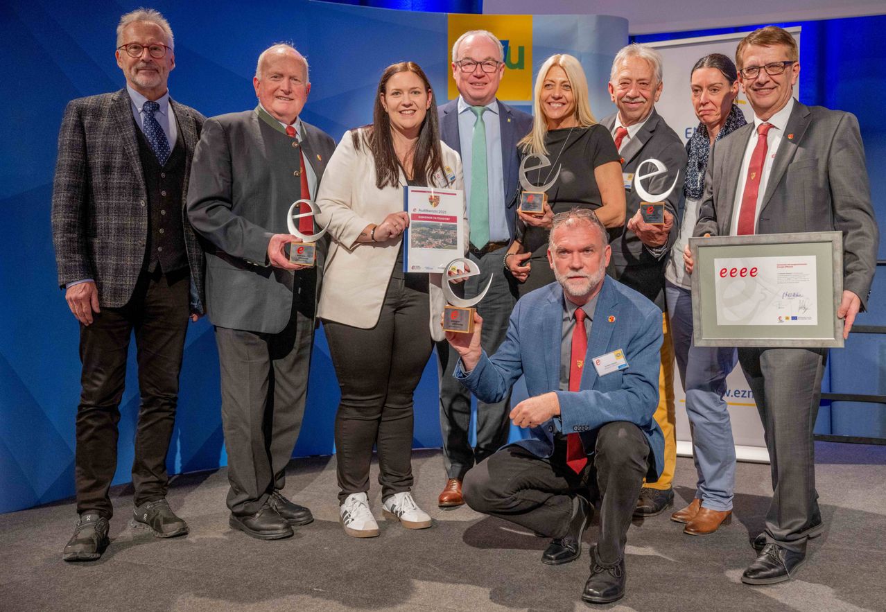 Eine Gruppe von Menschen in formeller Kleidung steht auf einer Bühne und hält Auszeichnungen. Ein Mann kniet vor ihnen und lächelt. Ein Banner mit Logos ist hinter ihnen.