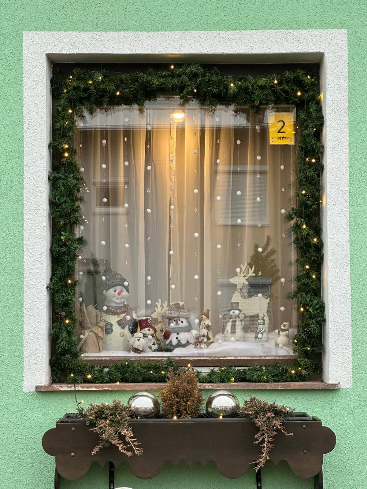 Ein Fenster, das mit festlichen Dekorationen für die Feiertage geschmückt ist. Der Fensterrahmen ist grün und mit einer Gardine verziert, die mit Lichtern und Ornamenten geschmückt ist. Darin befinden sich ein Schneemann und ein Rentier. Der Fenstersims hat zusätzliches Grün und dekorative Kugeln.