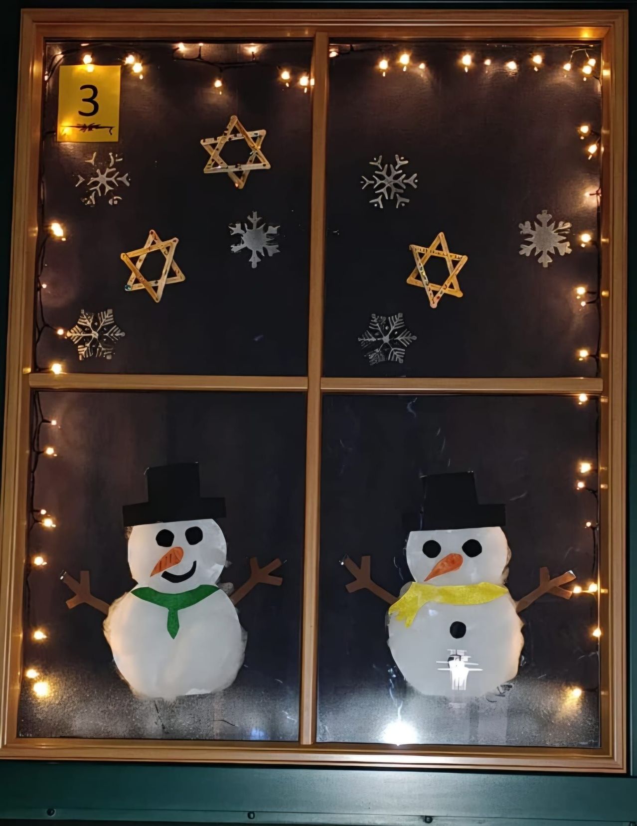 Zwei Schneemänner mit Mützen, Schals und lächelnden Gesichtern sind in einem Fenster ausgestellt. Sie sind von Schneeflocken und Davidsternen eingerahmt, wobei Lichterketten die Szene beleuchten.