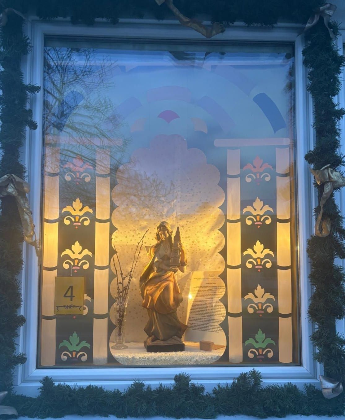 Ein Fensterdisplay zeigt eine goldene Statue einer Frau, die eine Laterne hält. Sie ist beleuchtet und von dekorativen Glaspaneelen mit Blumenmotiven umgeben. Eine Nummer 4 ist links sichtbar.