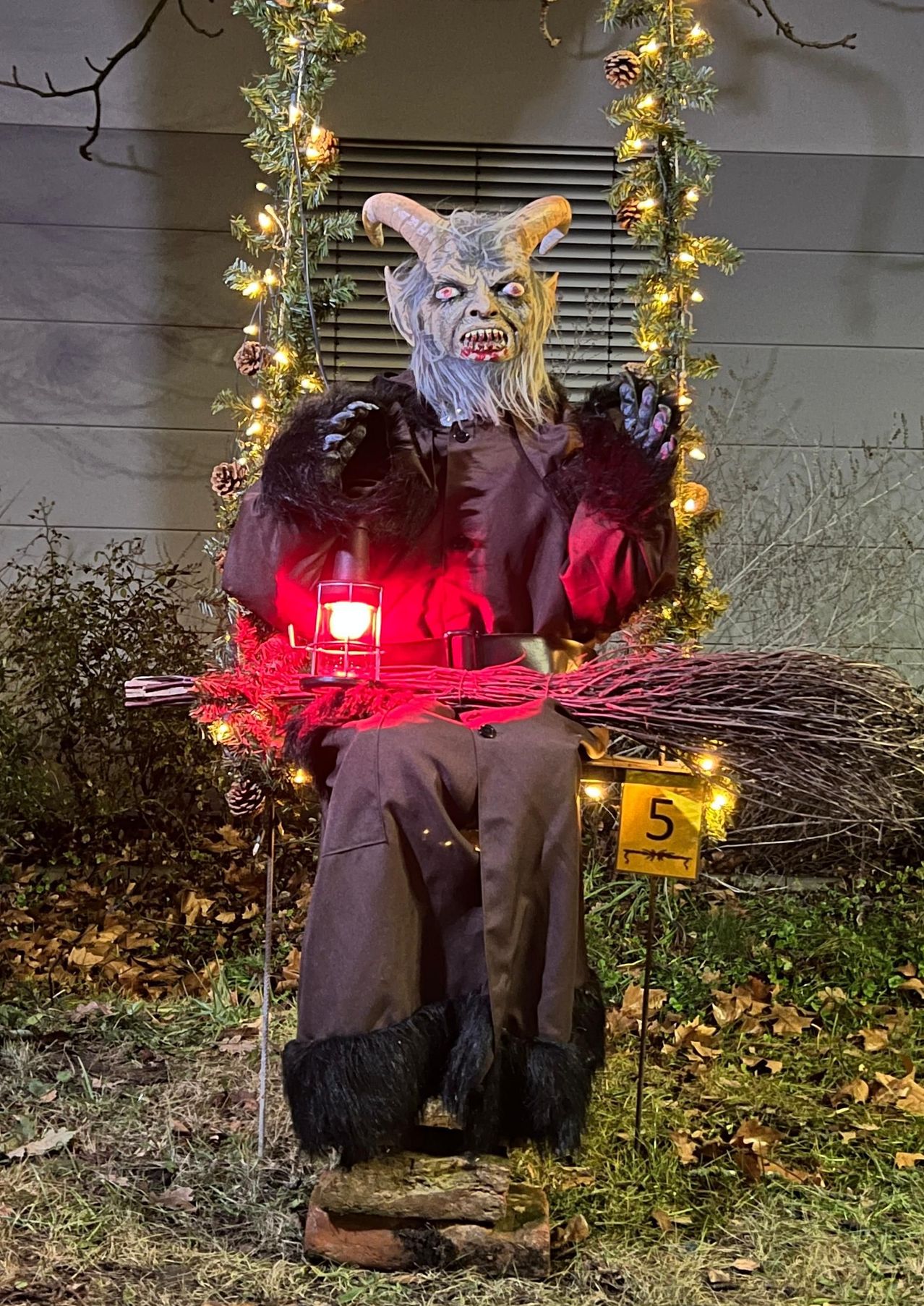 Eine Halloween-Dekoration eines Dämons sitzt auf einer Schaukel mit einer Laterne. Der Dämon hat Hörner, ein rotes Licht auf der Laterne und einen Besen vor sich.