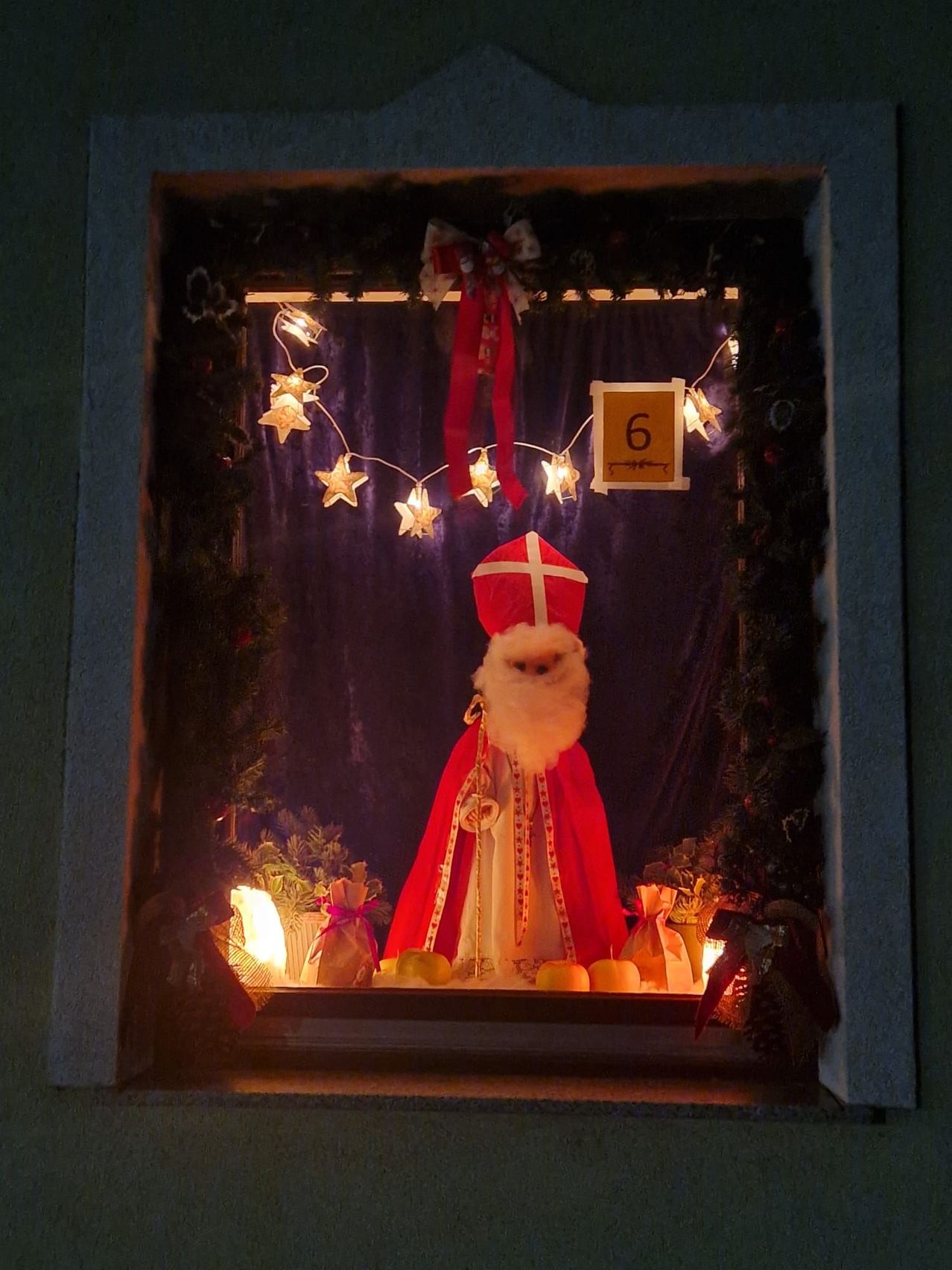 Ein Fensterdisplay zum sechsten Advent mit einer Figur des Weihnachtsmanns, einem roten Umhang und einem weißen Bart. Die Szene ist mit Weihnachtsdekorationen, Lichtern und einem Schild, das '6' zeigt, geschmückt.