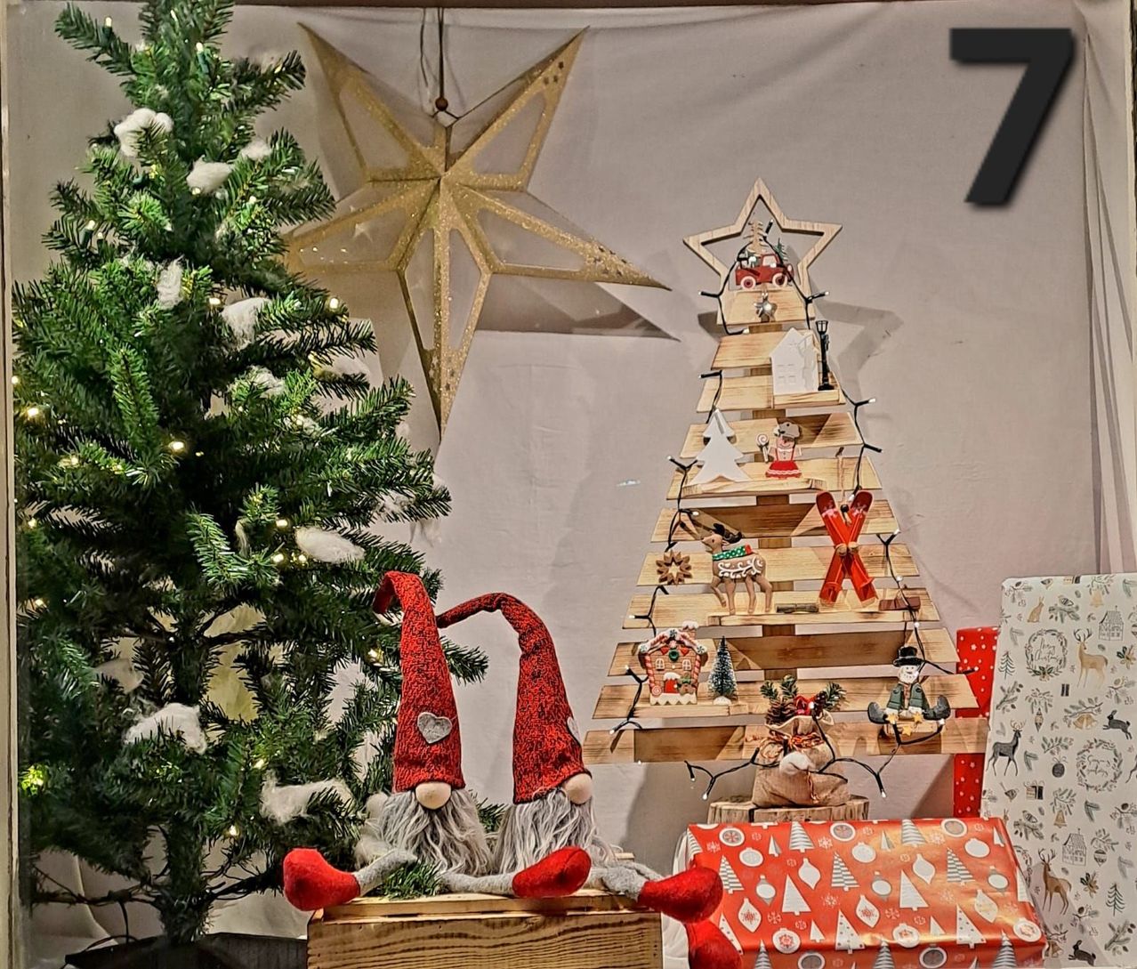 Einer hölzerne Weihnachtsbaum mit Lichtern und Ornamenten steht neben zwei Zwergen. Ein goldener Stern hängt an der Wand dahinter. Rechts ist ein weiterer Weihnachtsbaum aus Paletten und Geschenken ausgestellt.