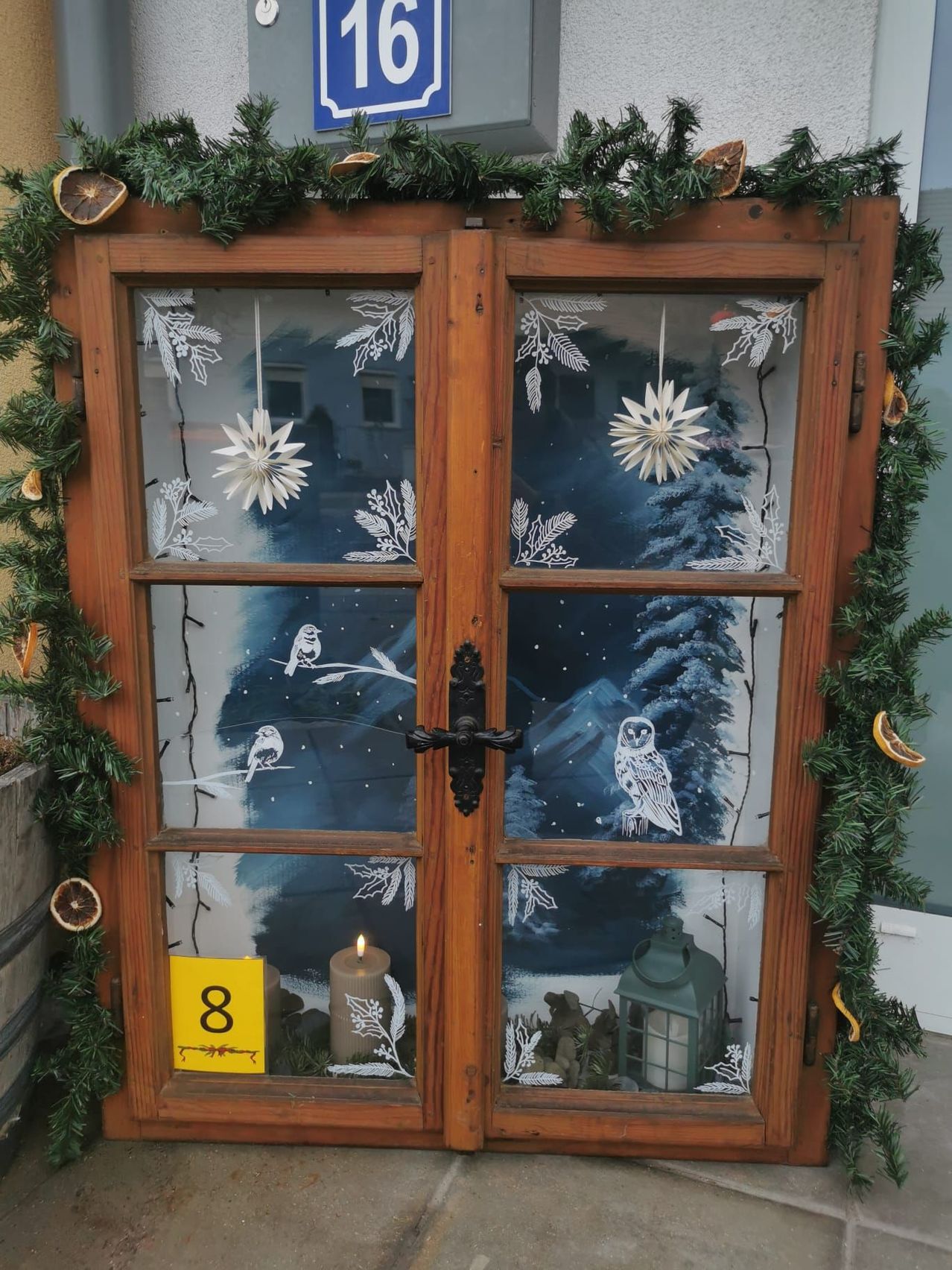 Ein hölzernes Fensterdisplay zeigt eine verschneite Szene mit einer Kerze, einer Eule und Vögeln auf einem Zweig. Schneeflocken und grüne Girlanden schmücken das Fenster. Ein gelbes Schild mit der Nummer 8 ist vorhanden.
