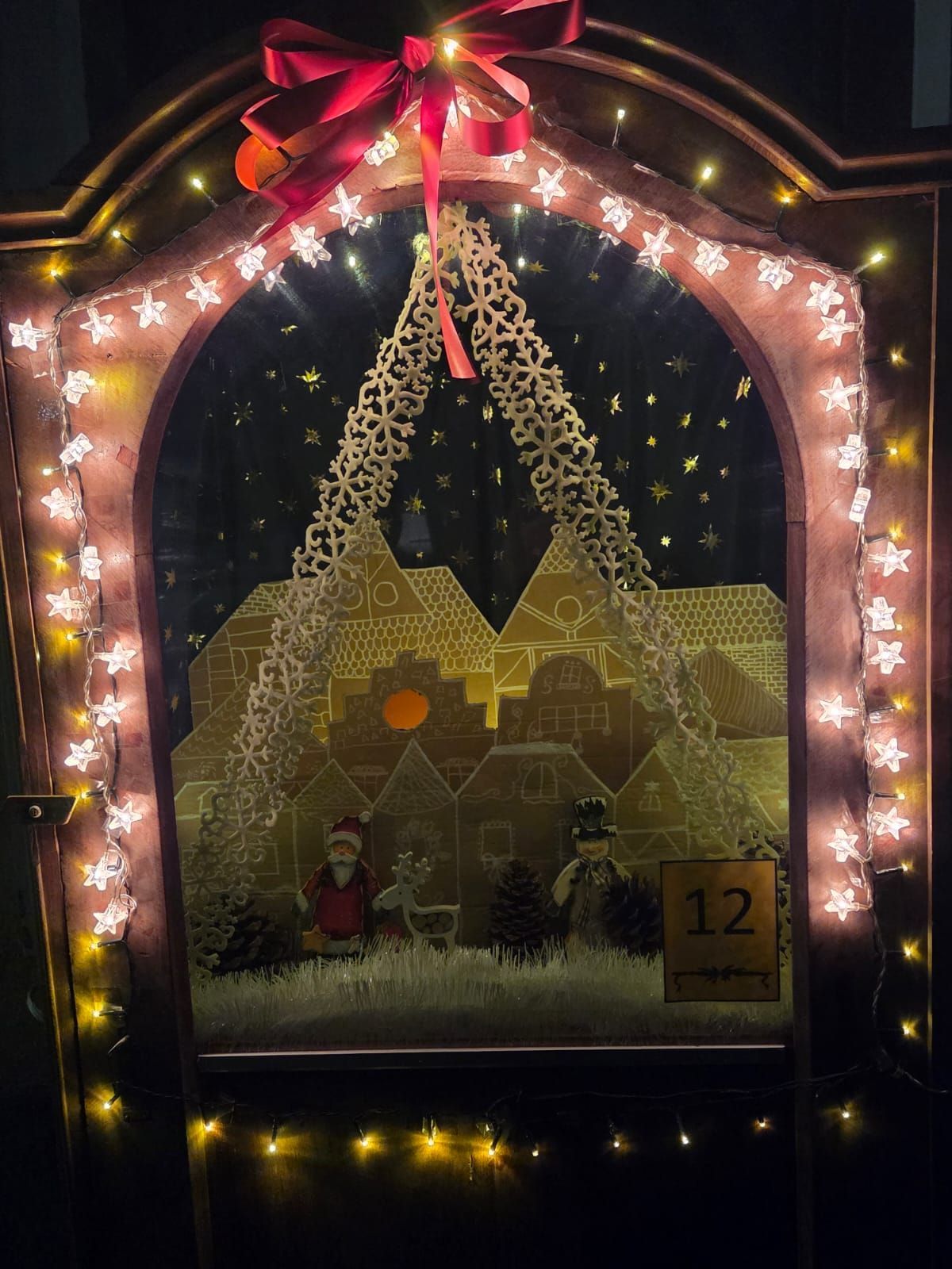 Ein Weihnachtsfenster mit einem Lebkuchenhaus, dem Weihnachtsmann und einem Rentier. Das Haus ist mit Lichtern und Sternen dekoriert und rechts steht die Zahl 12.