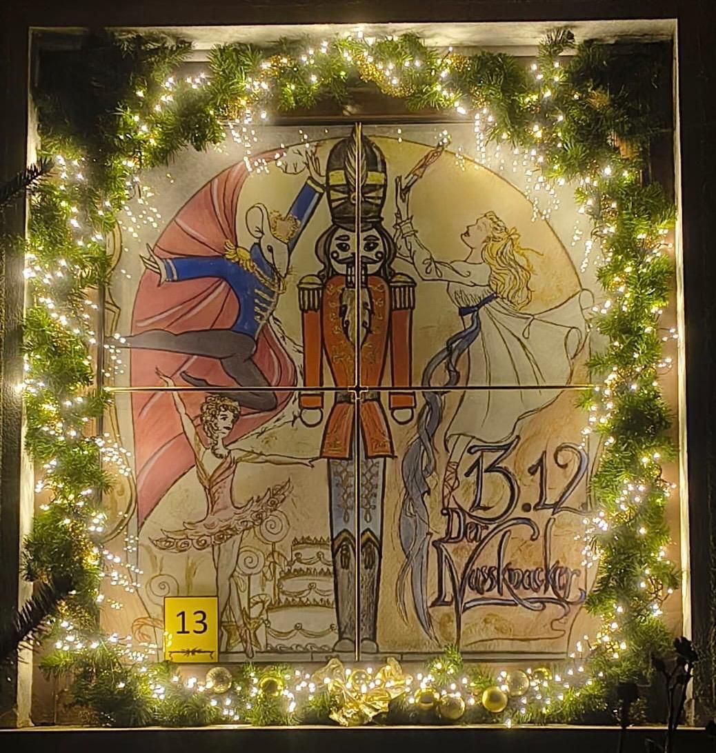 Ein Fensterdisplay mit einer Weihnachtsdekoration, die einen Nussknacker und Ballerinen zeigt. Die Nummer 13 befindet sich in einem gelben Etikett unten links. Der Rahmen ist von einem grünen Kranz mit weißen Lichtern umgeben.