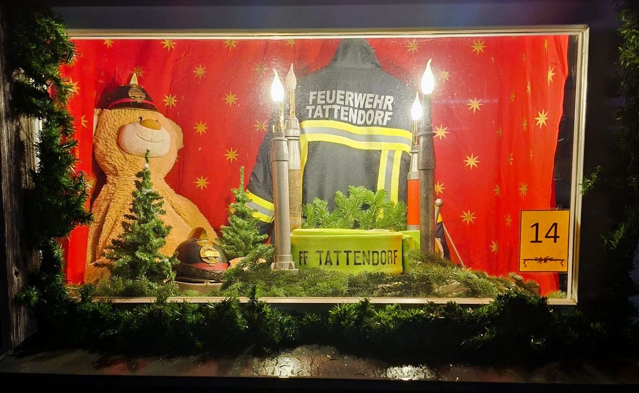 Eine festliche Präsentation mit einem Feuerwehrmann-Jacke, auf der 'Feuerwehr Tattendorf' steht, einem Teddybär, einem Feuerwehrhelm und einer gelben Tasse mit dem Text 'FF Tattendorf'. Der Hintergrund ist rot mit gelben Sternen.