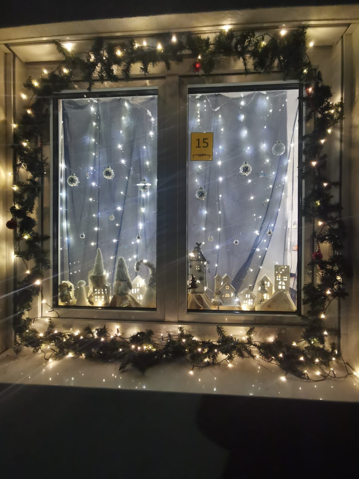 Zwei Fenster zeigen eine Weihnachtsdorfszene mit Gebäuden, Zwergen und Lichtern. Festliche Dekorationen und Weihnachtskugeln schmücken die Fensterbänke. Eine Nummer 15 ist auf einem Fenster sichtbar.
