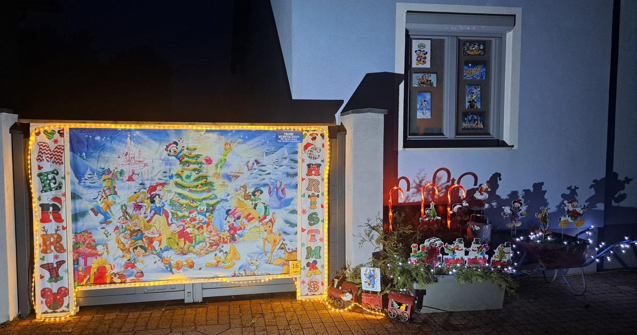 Ein Disney-Weihnachtsrätsel an der Wand eines Hauses. Es ist mit gelben Lichtern beleuchtet. Auf der rechten Seite befinden sich zwei kleine Weihnachtszüge. Das Fenster zeigt Weihnachtskarten.