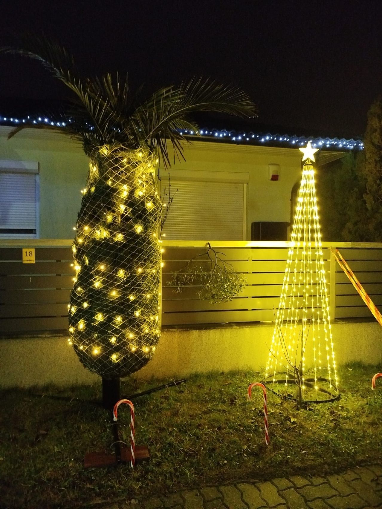 Ein Ananas mit gelben LED-Lichtern steht neben einem kleinen, beleuchteten Weihnachtsbaum vor einem Haus mit blau beleuchtetem Dach und einem Fenster. Der Boden ist mit Gras bedeckt.