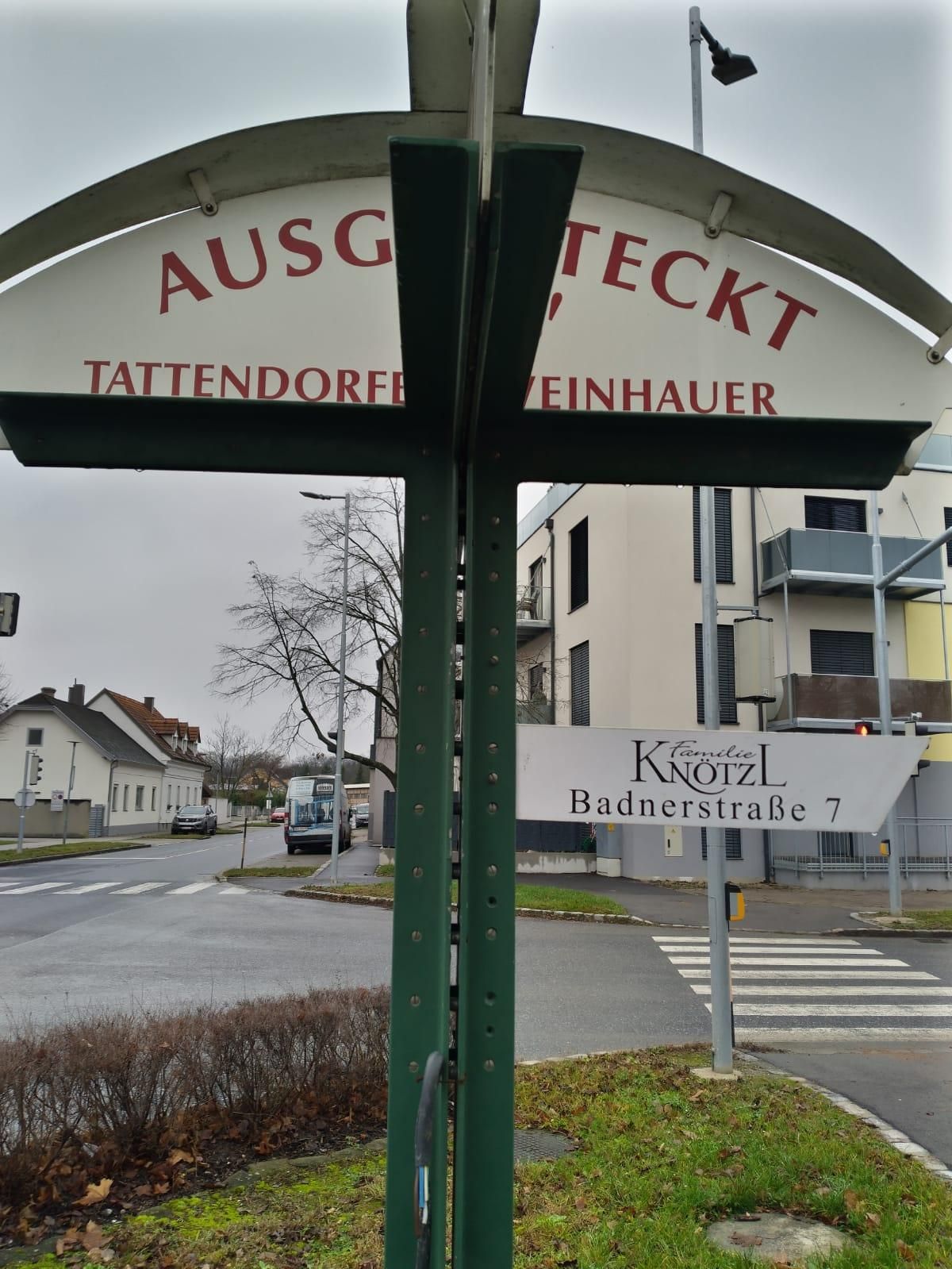 Ein Straßenschild an einer Kreuzung zeigt 'Ausg, TECKT Tattendorf Einhauer'. Ein Familiennamensschild zeigt 'Familie KNOTZL Badnerstraße 7'. Im Hintergrund sind Gebäude, Autos und ein Bus zu sehen.