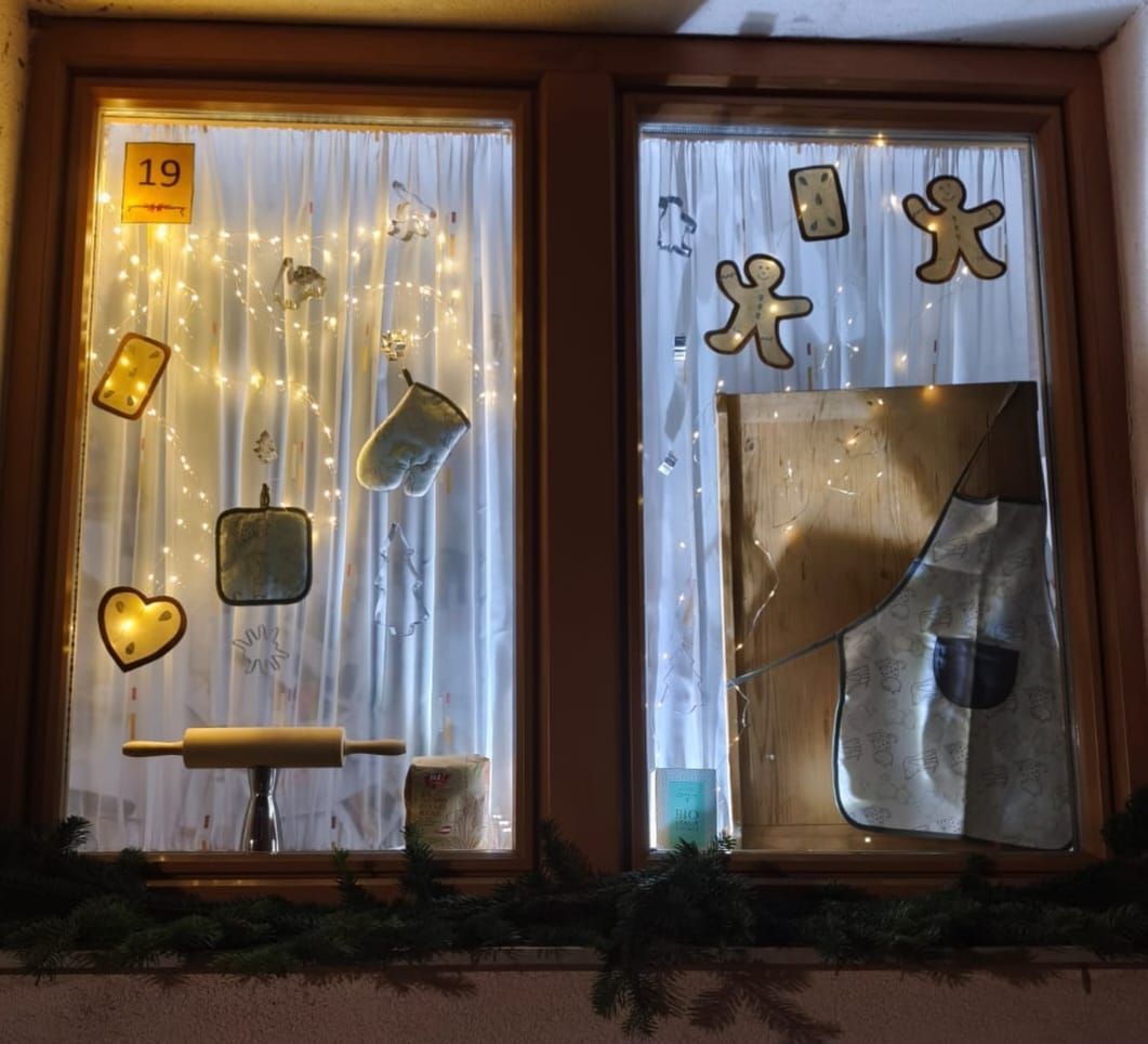 Zwei Fenster, die mit Lebkuchenmännern, Keksen, Ofenhandschuhen und einem Nudelholz dekoriert sind. Jedes Fenster hat die aufleuchtende Nummer 19. Ein Fenster hat ein weißes Küchentuch hängend.