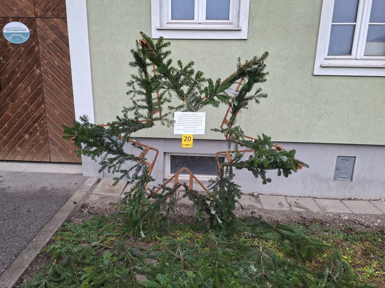 Ein sternförmiges Gebilde aus immergrünen Zweigen steht vor einem Gebäude mit Fenstern und einem gelben Schild mit der Nummer 20.
