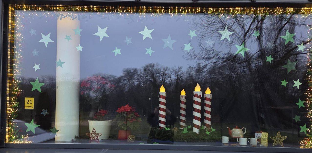 Eine Anzeige mit vier beleuchteten gestreiften Kerzen und roten Blumen in einem Fenster mit Weihnachtsthema. Draußen vor dem Fenster sind Bäume und eine Reflexion eines Gebäudes.