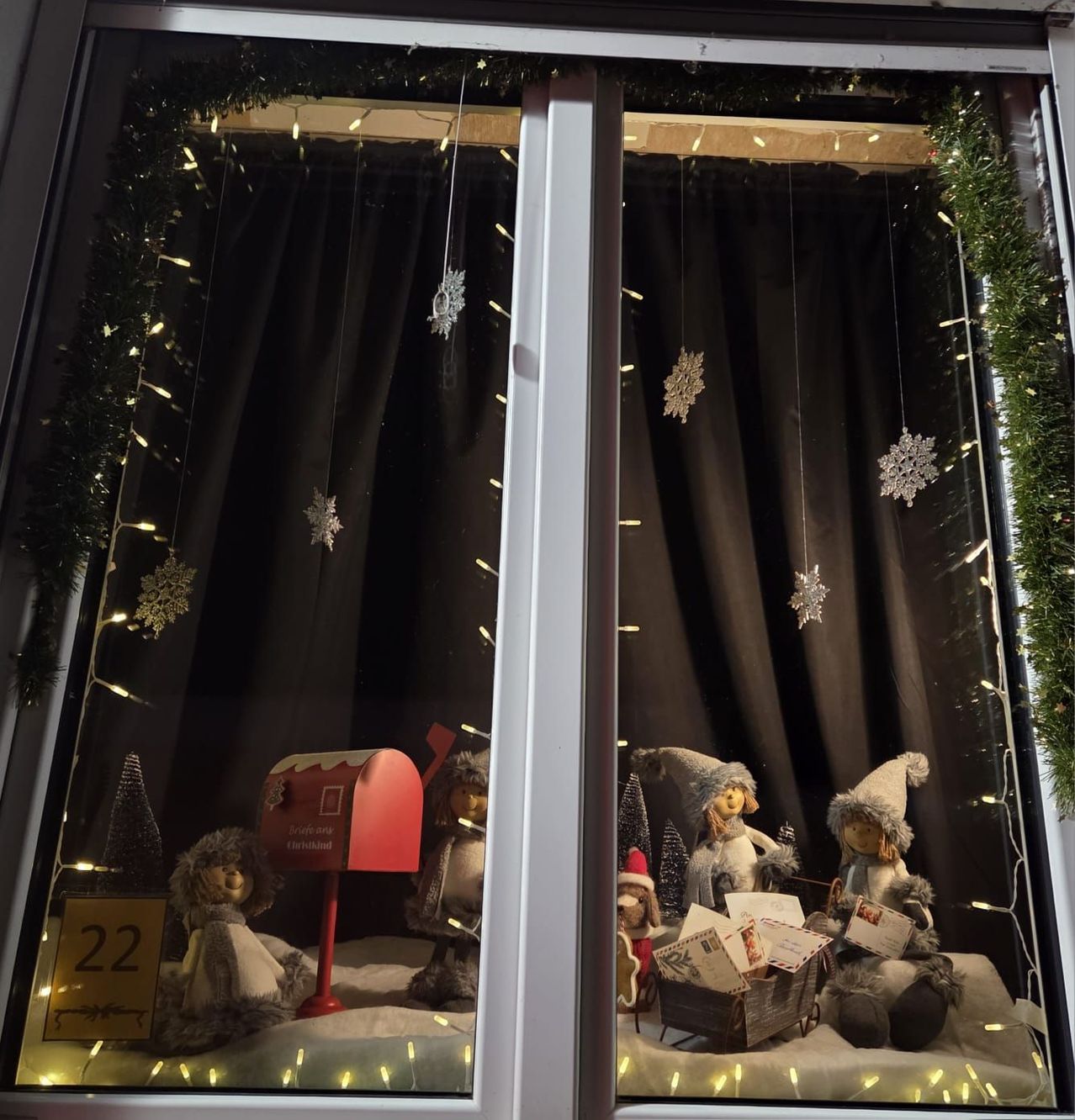 Eine Weihnachtsdekoration in einem Fenster mit einem roten Briefkasten, Puppen und Weihnachtsbäumen. Schneeflocken und Lichter schmücken das Fenster und schaffen eine festliche Atmosphäre.