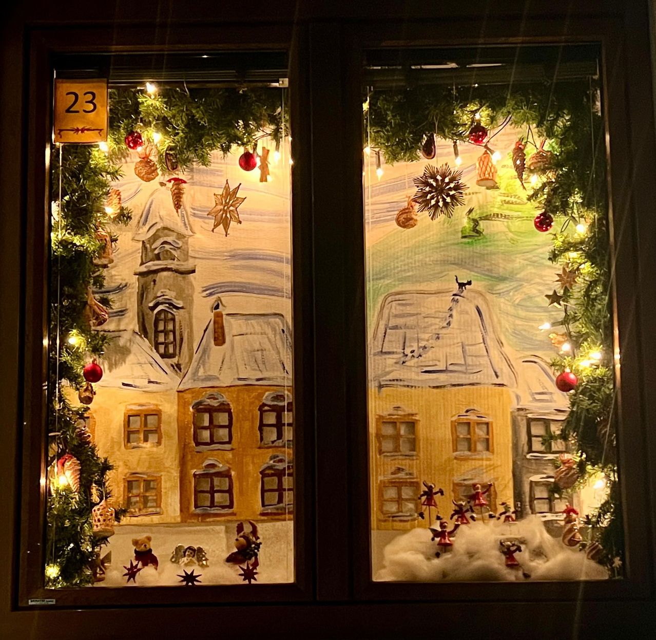 Zwei Fenster zeigen festliche Winterszenen mit Weihnachtsdekorationen. Beide haben eine gemalte Stadtszene mit Gebäuden, einer verschneiten Landschaft und Weihnachtslichtern. Die Nummer 23 befindet sich am linken Fenster.