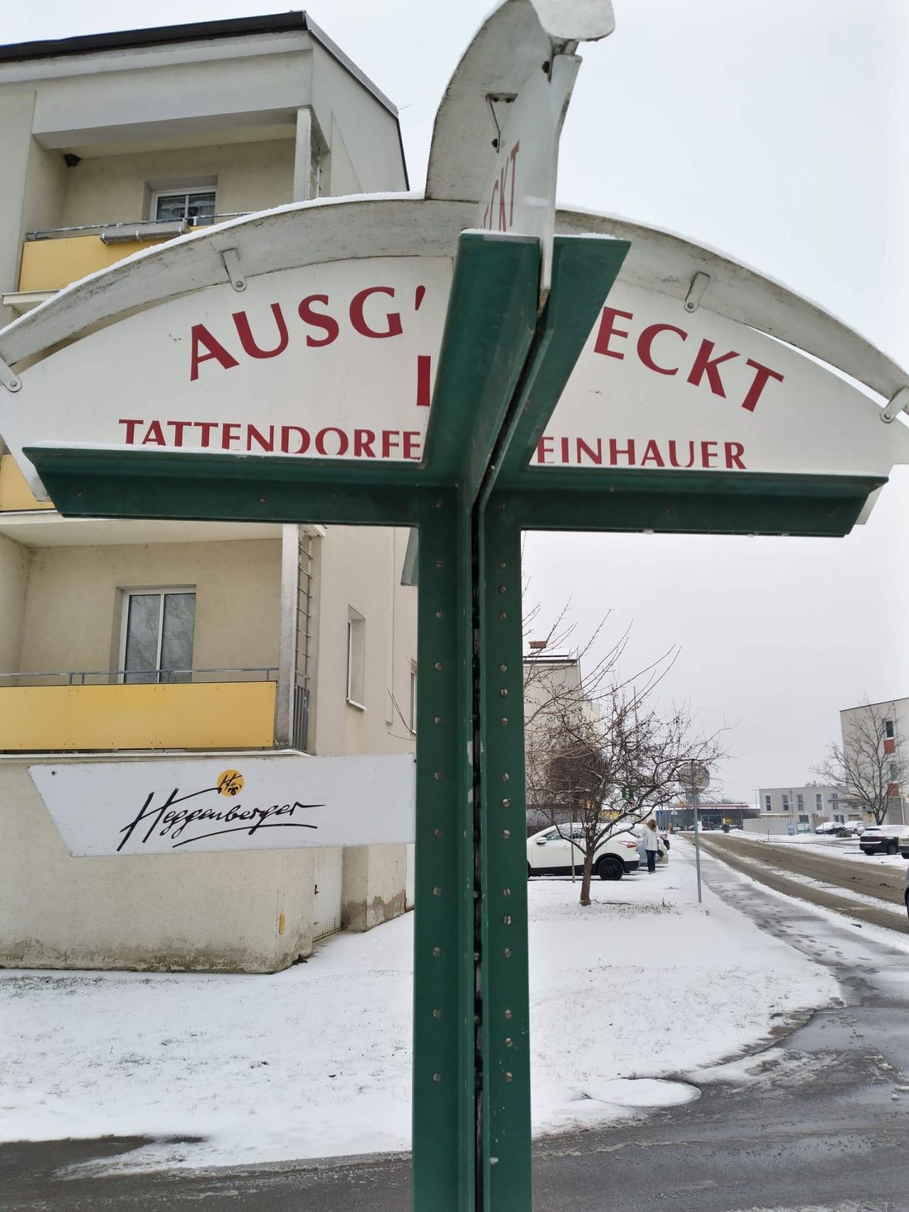 Ein Straßenschild mit dem Namen 'AUSG'ECKT TATTENDORFE FINHAUER' steht vor einem Gebäude in einem verschneiten Gebiet. Ein Schild darunter lautet 'Huggerburger'. Ein weißes Auto ist auf der Seite geparkt. Bäume und andere Gebäude sind im Hintergrund.