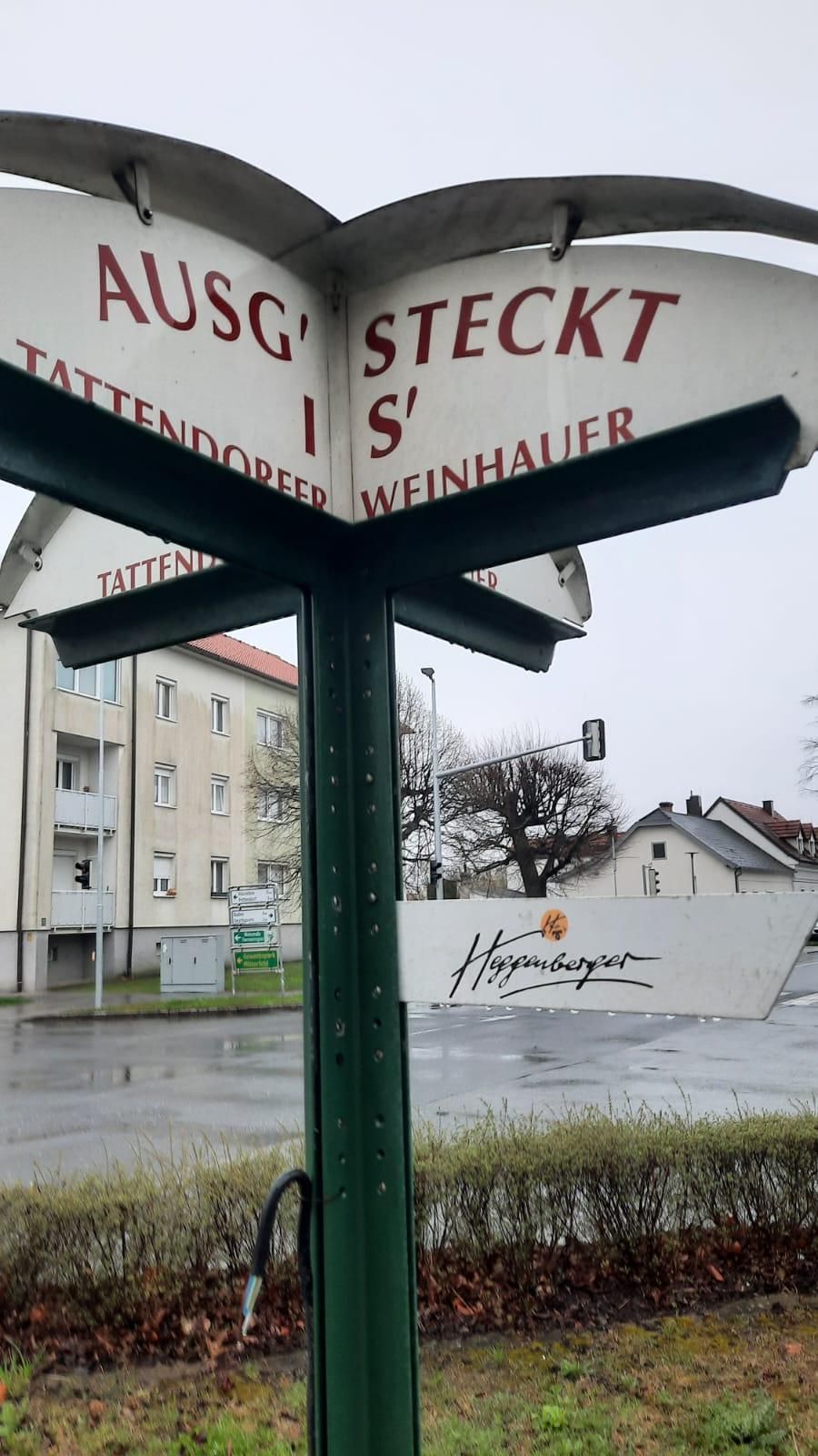 Ein Schild für Tattendorfer Weinhauer steht im Regen an einem Pfosten. Ein Gebäude mit Balkonen befindet sich hinter dem Schild.