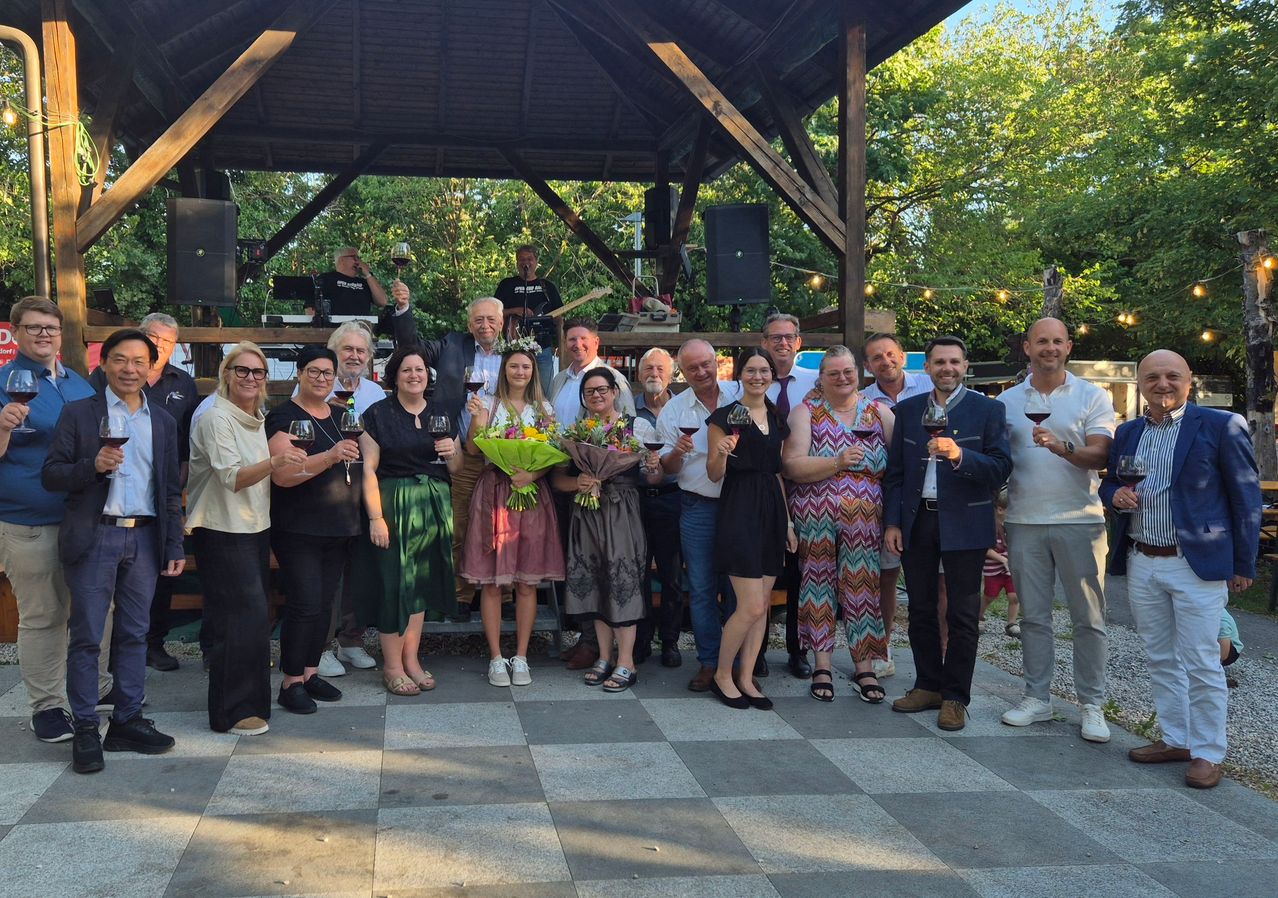 Eine Gruppe von Menschen posiert mit Weinglas, Blumen und Blumensträußen für ein Foto. Dahinter ein Pavillon mit Lautsprechern und einem Gitarristen.