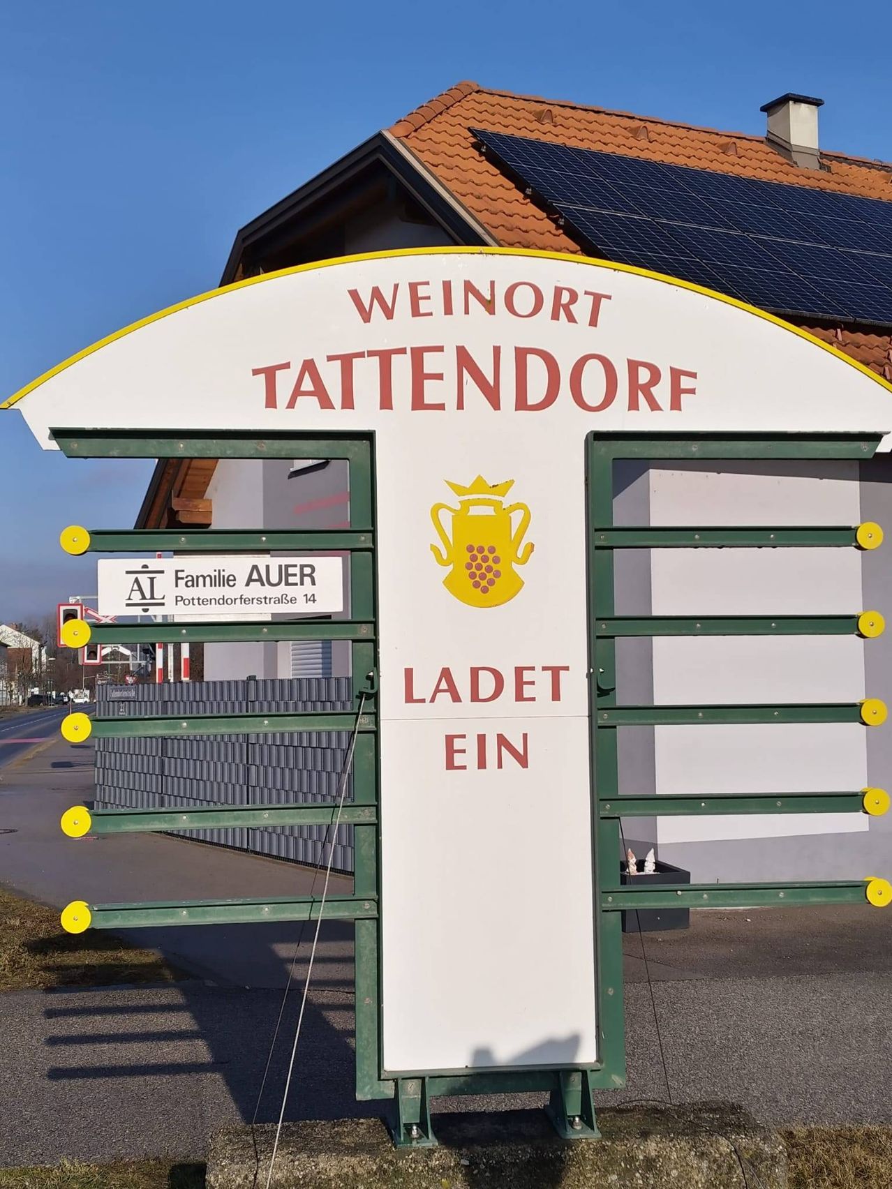 Ein Schild für Weinort Tattendorf steht vor einem Gebäude. Das Schild zeigt einen goldenen Pokal und den Text "LADET EIN". Ein kleineres Schild darunter trägt den Schriftzug 'Familie AUER'.