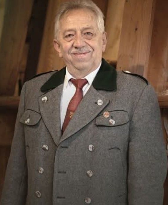 Ein älterer Mann mit einem Schnurrbart, der einen grauen Mantel mit einer roten Krawatte und mehreren Knöpfen trägt, lächelt und steht vor einer Holzwand.