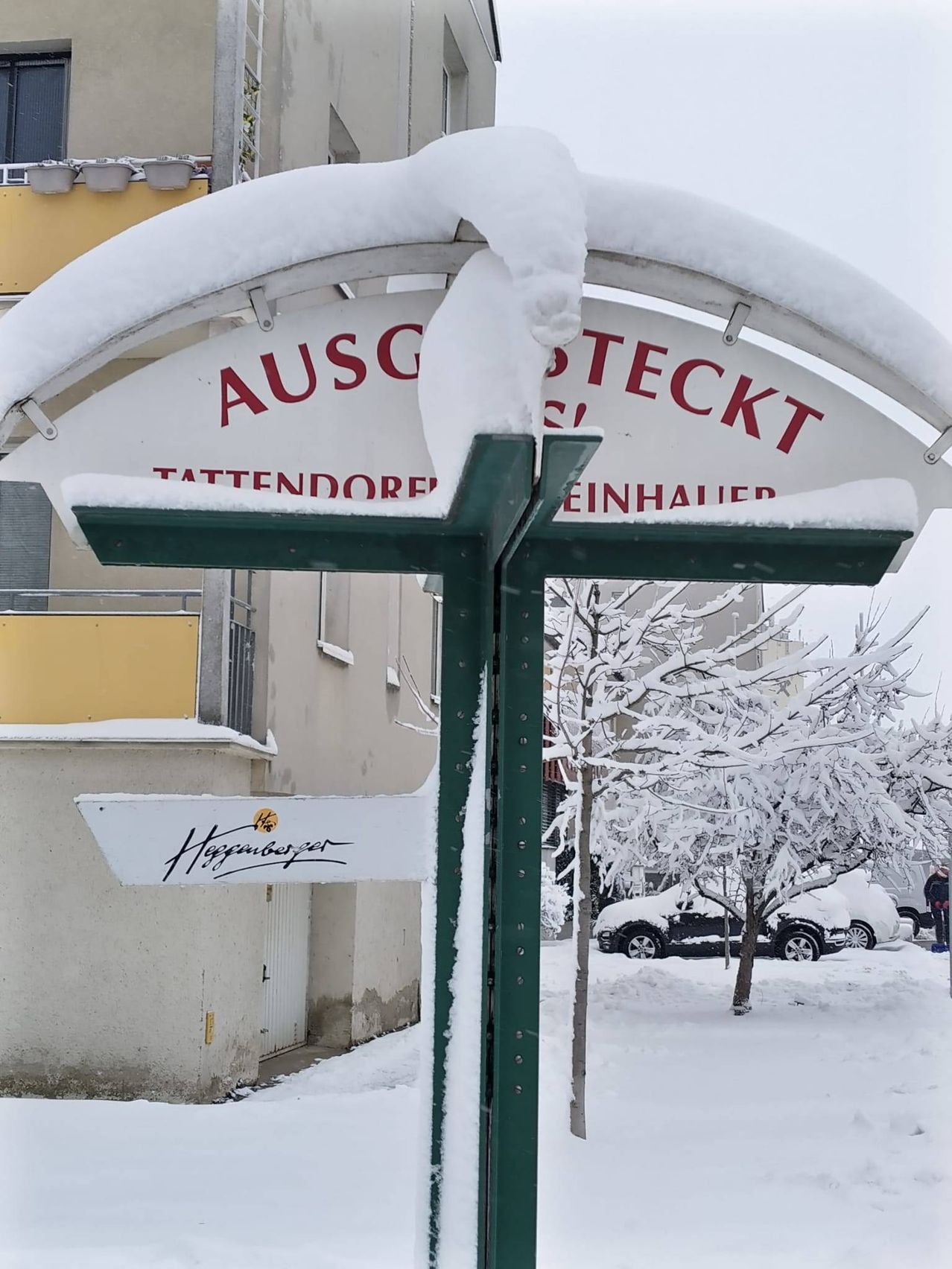 Ein verschneiter Straßenschild für Ausstekkt Tattendorf Einh steht neben einem Gebäude. Dahinter sind mit Schnee bedeckte Bäume. Vor dem Gebäude ist ein Auto geparkt.